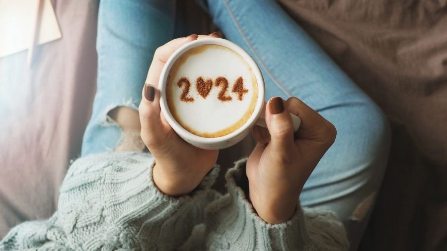 New year 2024 celebrated coffee cup with number 2024 on frothy surface of cappuccino in coffee cup holding by woman in green knitted sweater with jeans sitting on brown bed with notebook and pencil.